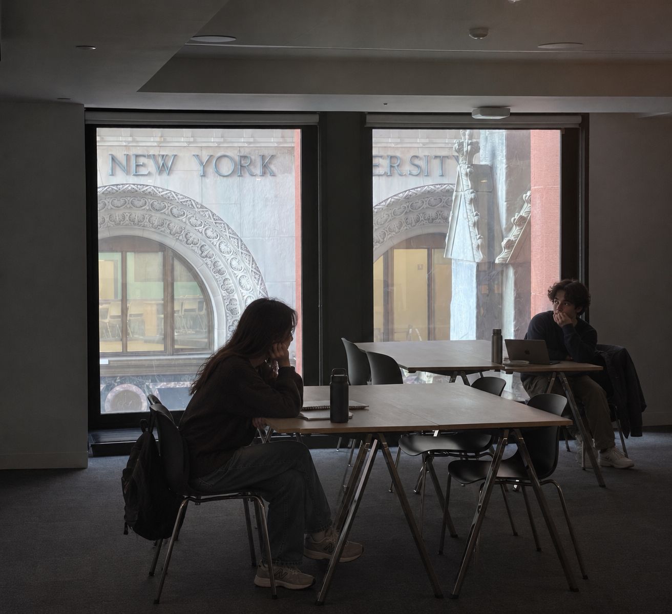 Two students seated at separate tables, with New York University visible through the windows.